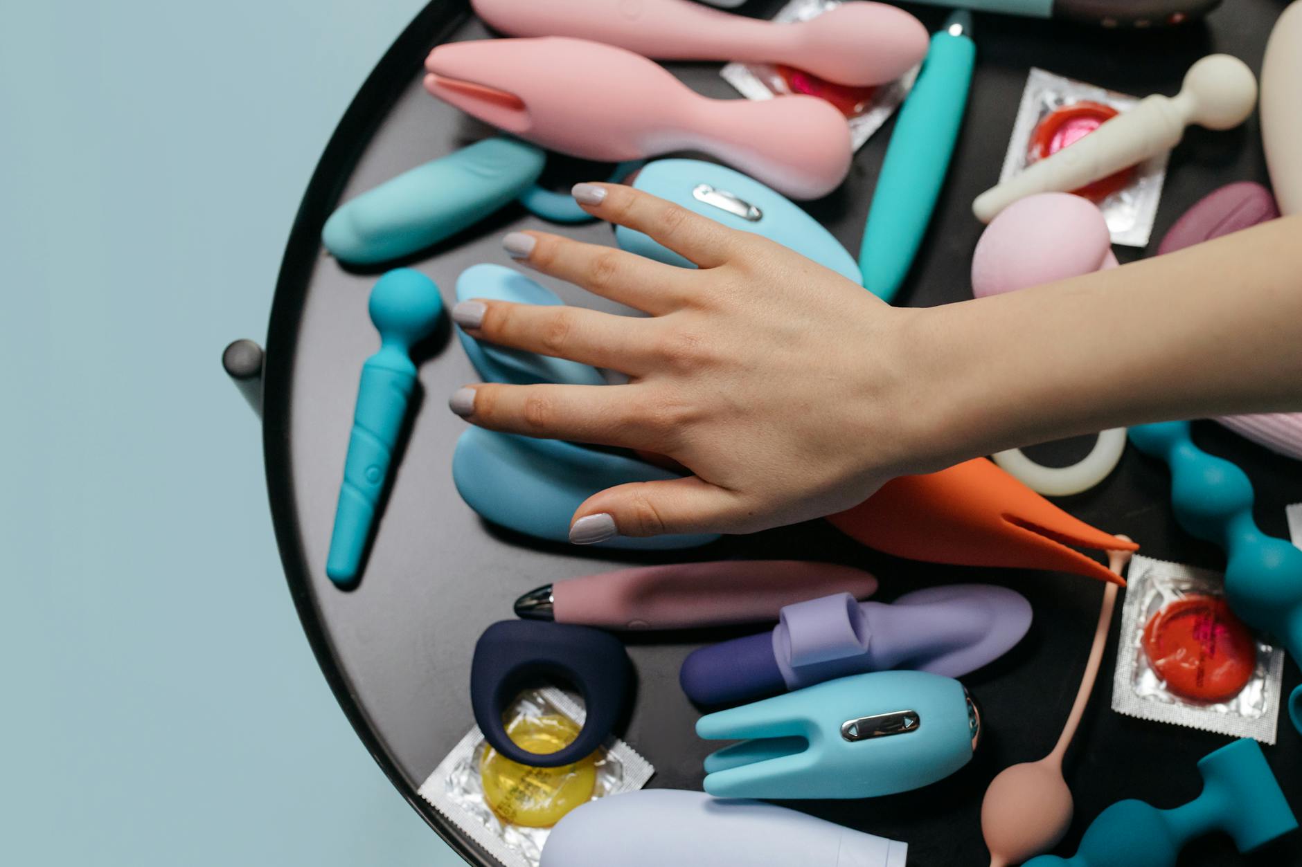 A hand reaching over a variety of colorful sex toys arranged on a table, representing openness and choice in intimate wellness.