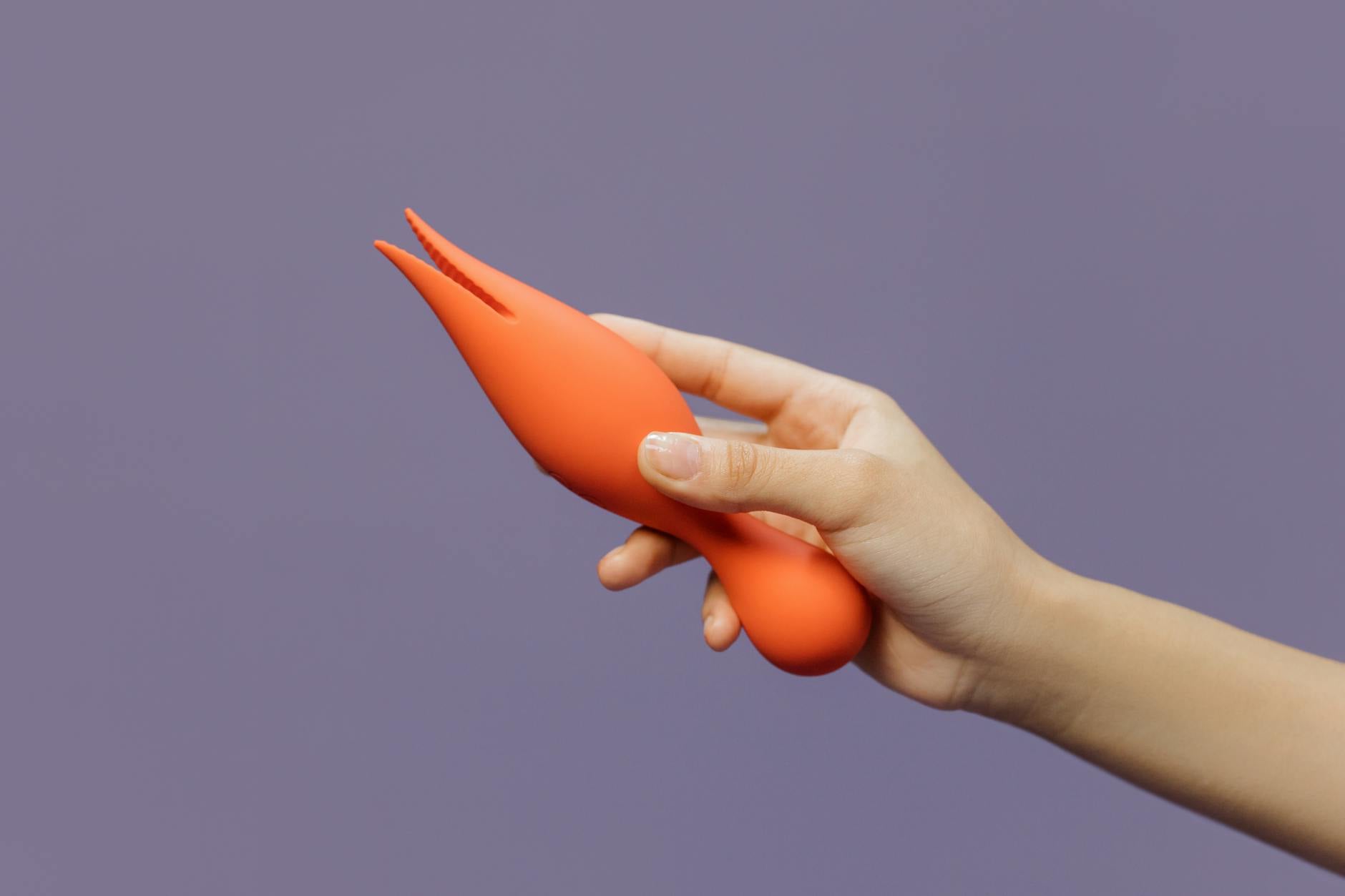 A hand holding a bright yellow clitoral vibrator against a minimalist purple backdrop