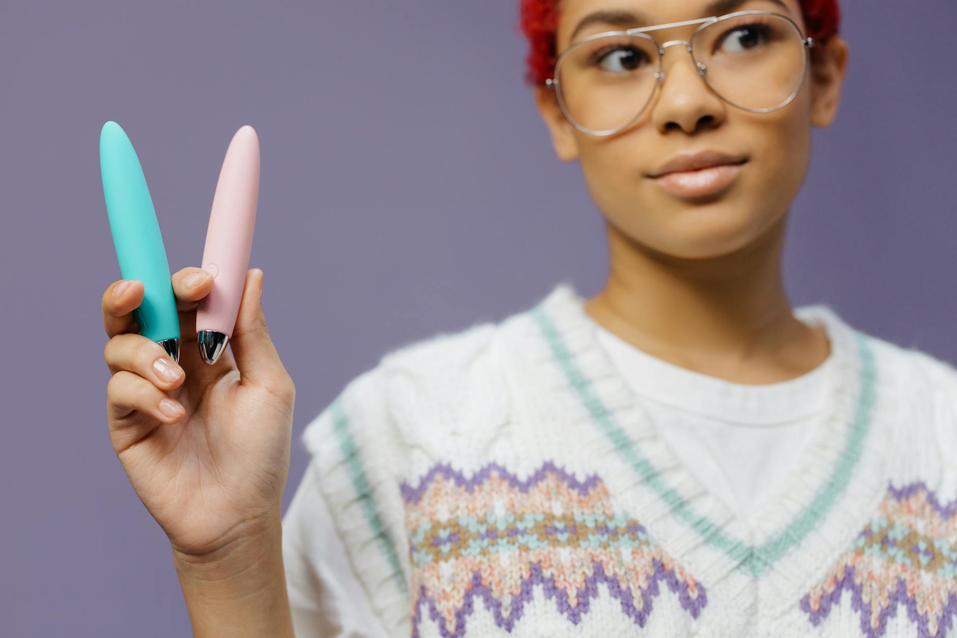 Two hands holding pink and blue silicone vibrators against a pastel background