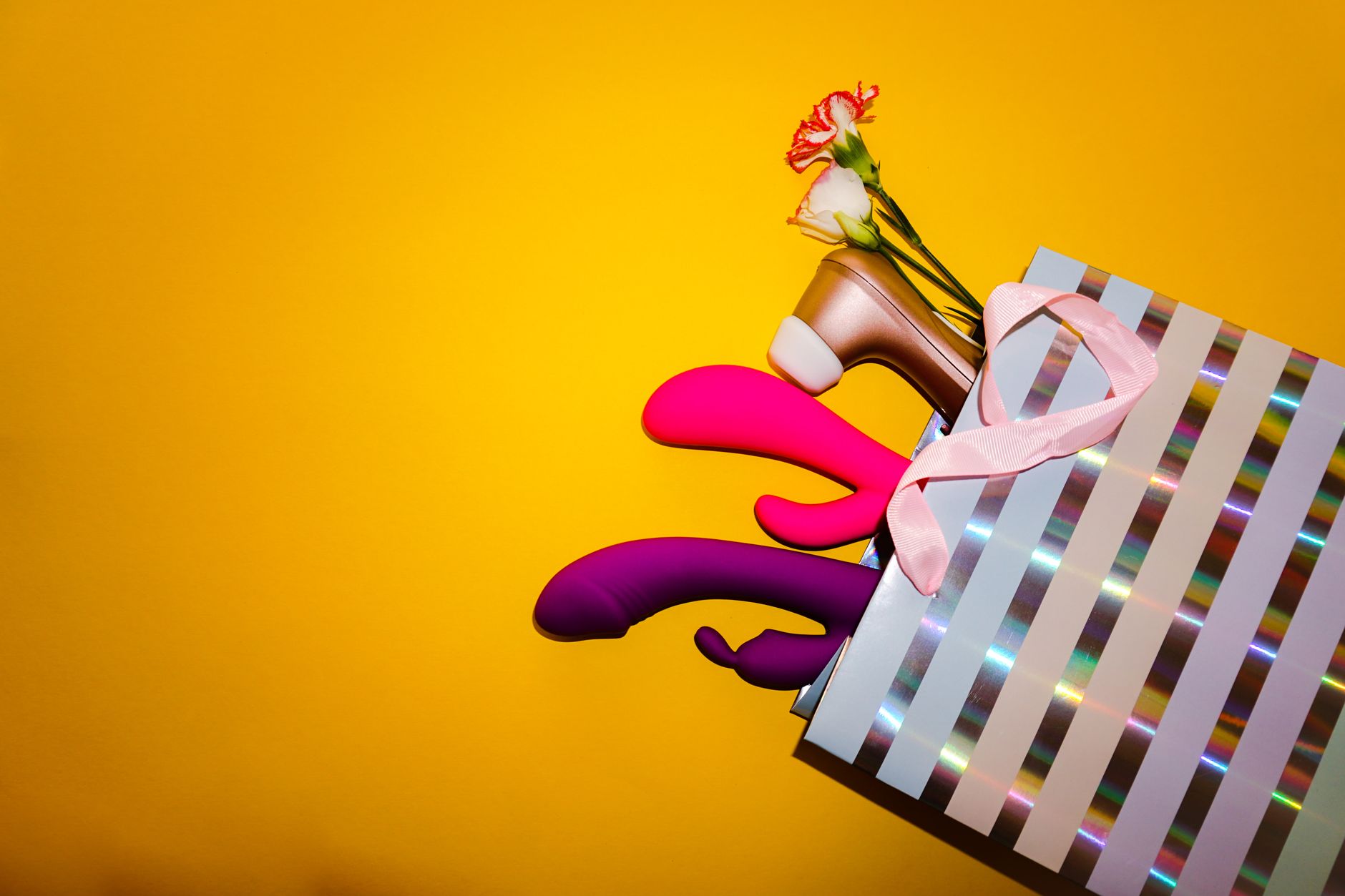 Colorful vibrators arranged with flowers in a holographic gift bag against a bold yellow background.